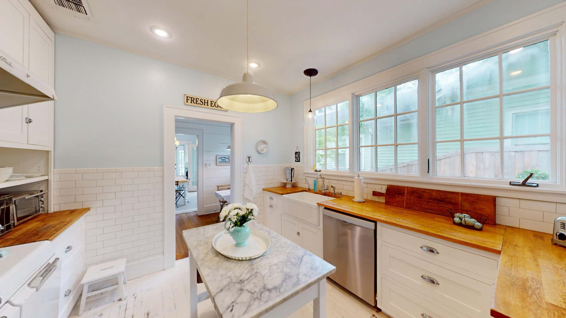 Kitchen from the opposite angle showing farmhouse sink, marble-topped island, butcher-block counters, and a wall of windows over the backyard