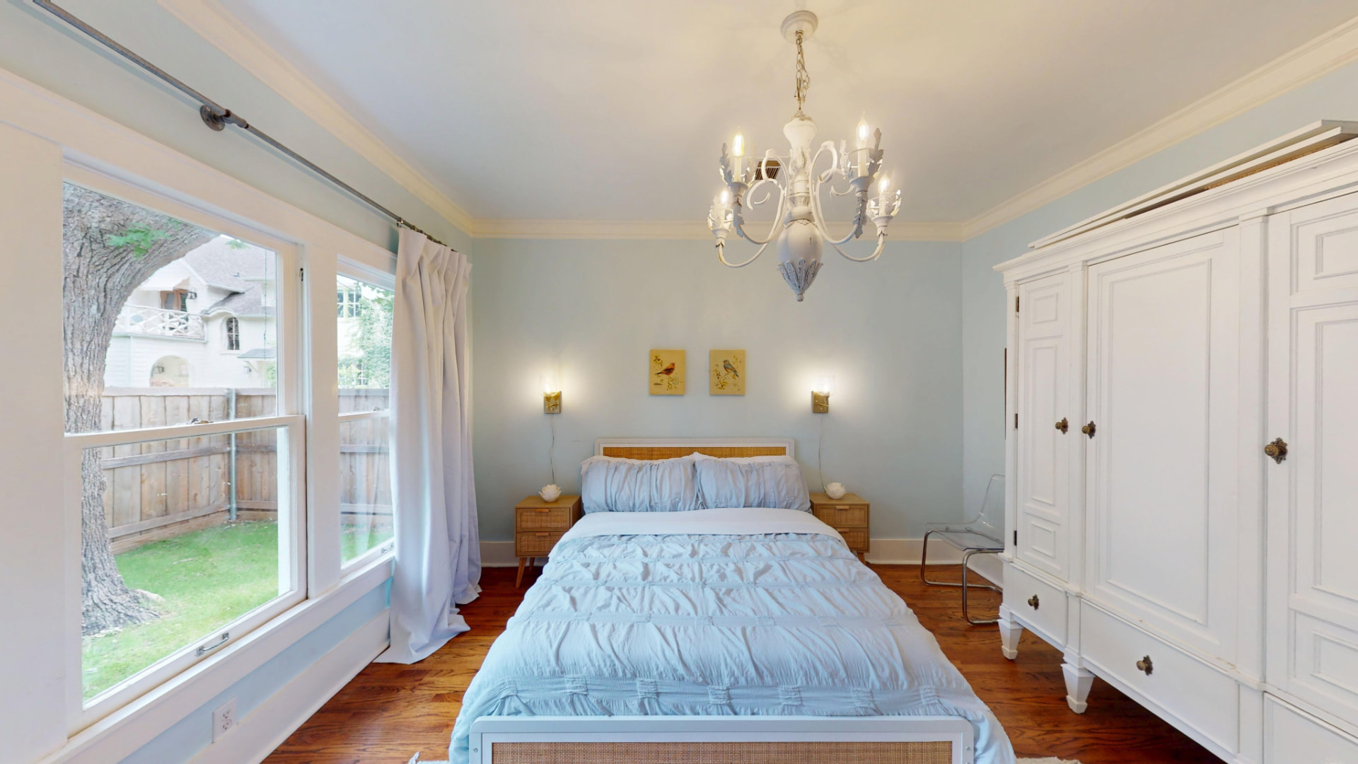 Guest bedroom with pale blue walls, vintage chandelier, cane bed, and a tall window overlooking the side garden