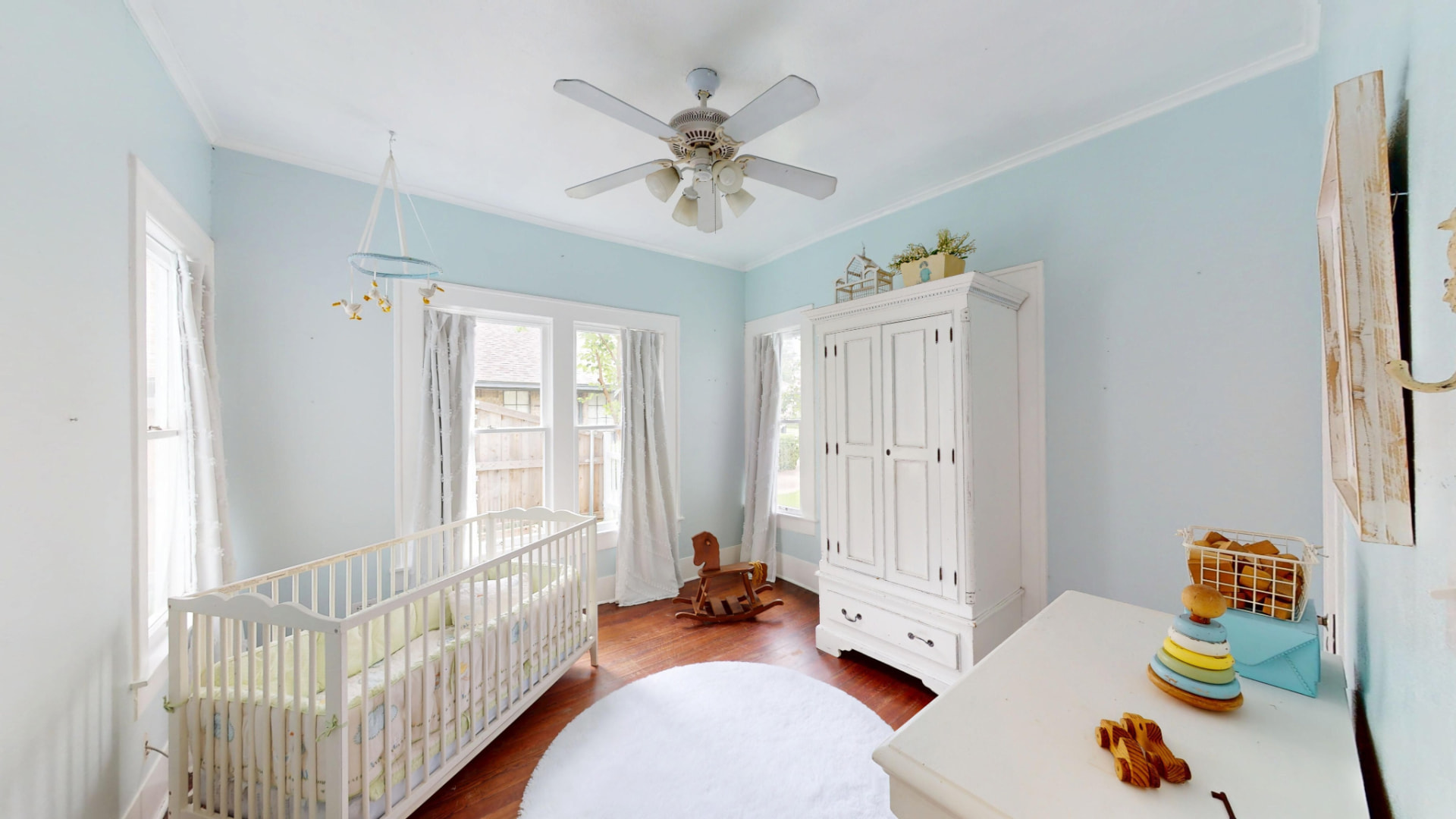 Nursery with pale blue walls, white crib, vintage armoire, and a small rocking horse on hardwood floors