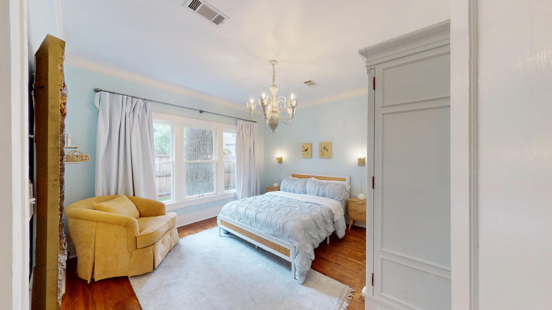 Bedroom with pale blue walls, chandelier, mustard-yellow armchair, and a queen bed beneath tall windows