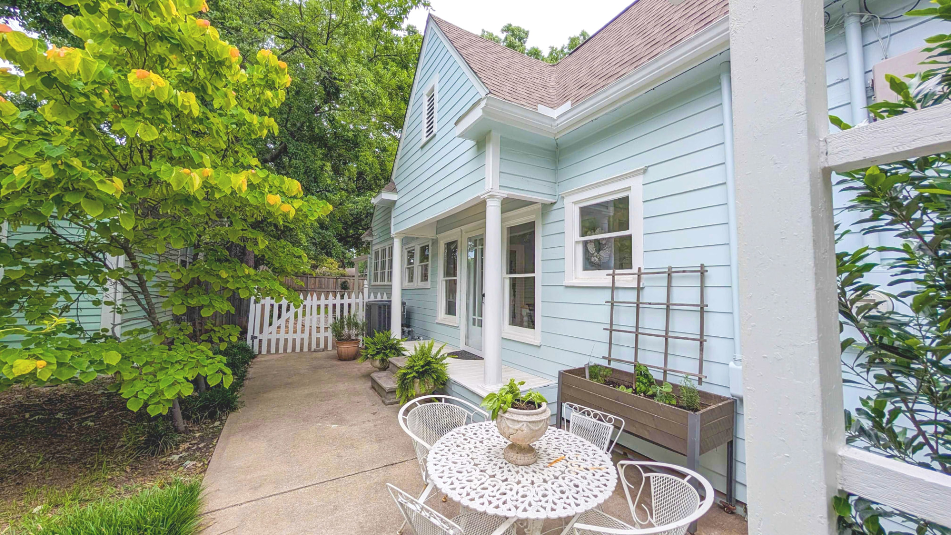Sea-glass blue side elevation with a small patio, wrought-iron bistro set, redbud tree, and white picket gate to the backyard